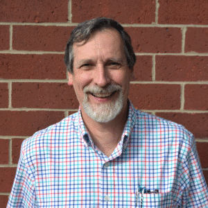 Photo of Paul Levy wearing a plain shirt and standing in front of a brick wall.