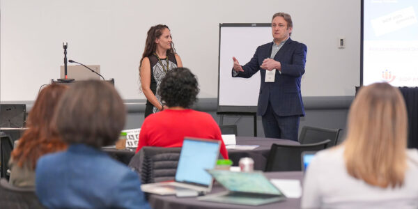 A man and woman stand together in front of a large screen, presenting a presentation.