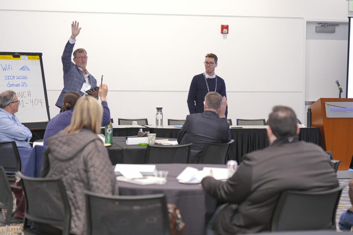 Attendees sitting at tables in a conference room, participating in a workshop. While speaker is talking in front of room with hand raised.