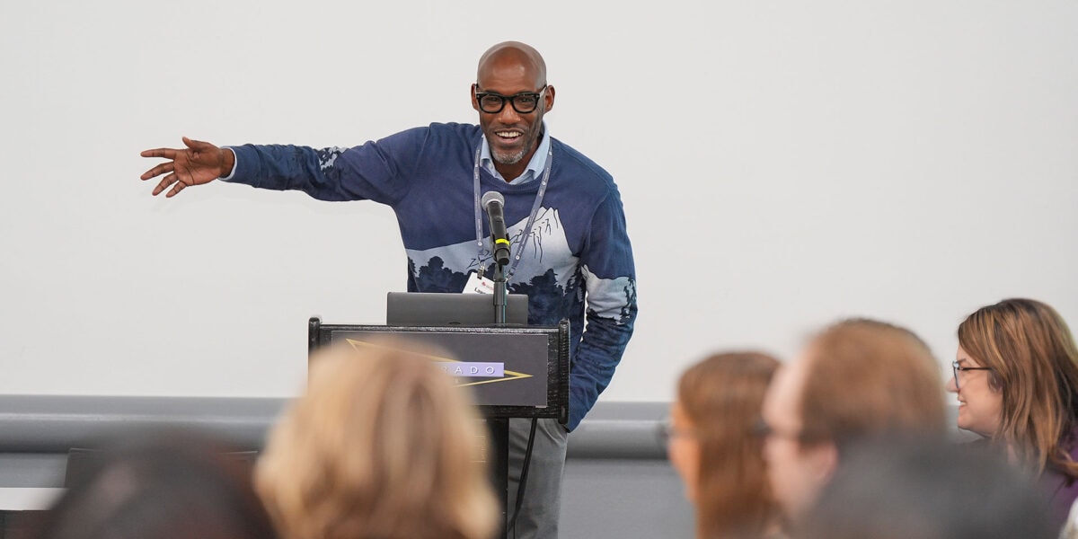 A man in glasses stands before an audience, giving a presentation and interacting with the attendees.