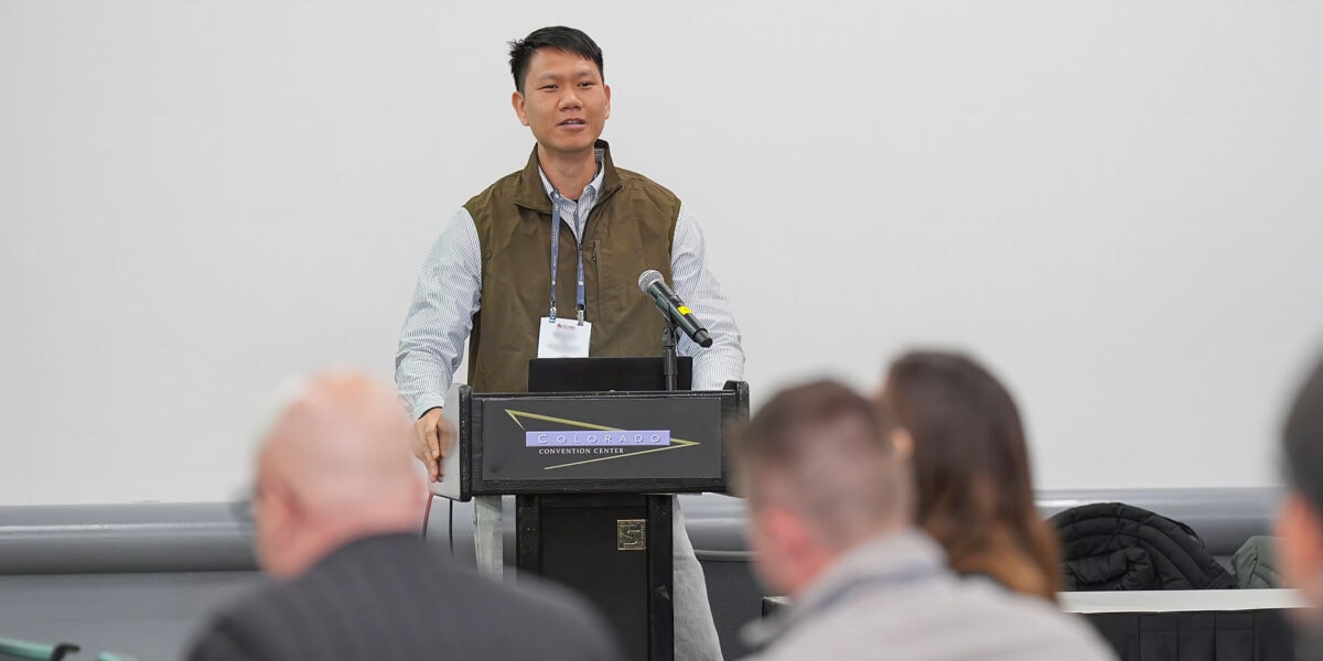 A man speaking at a conference, engaging the audience while standing next to a display screen with presentation materials.