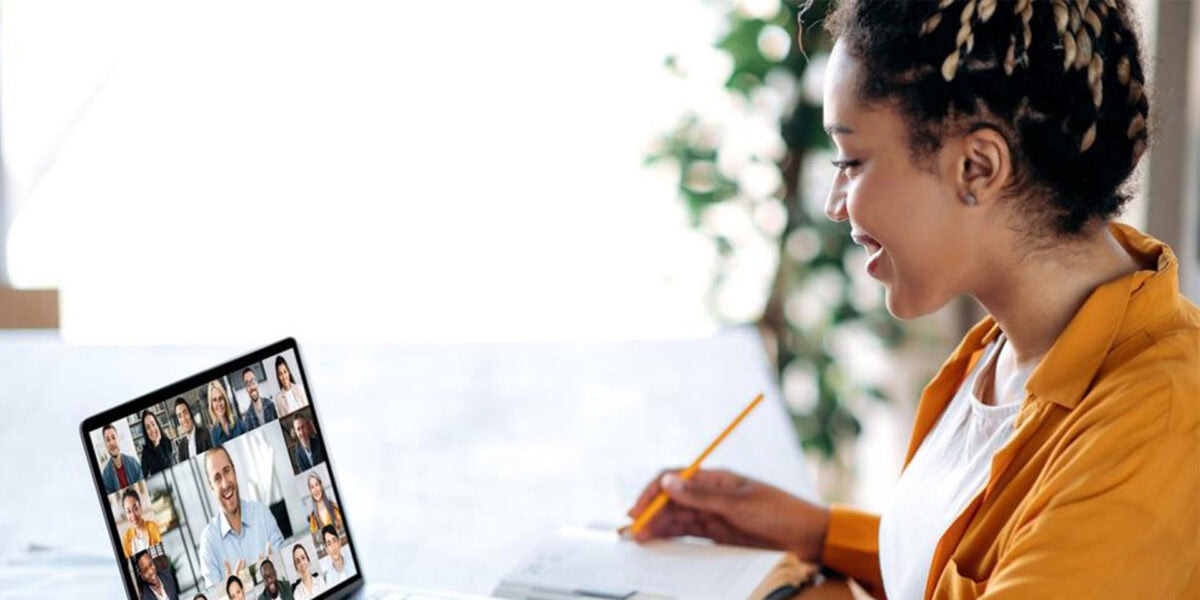 A woman sits at a desk with a laptop open and a pen in hand, attending a video call.
