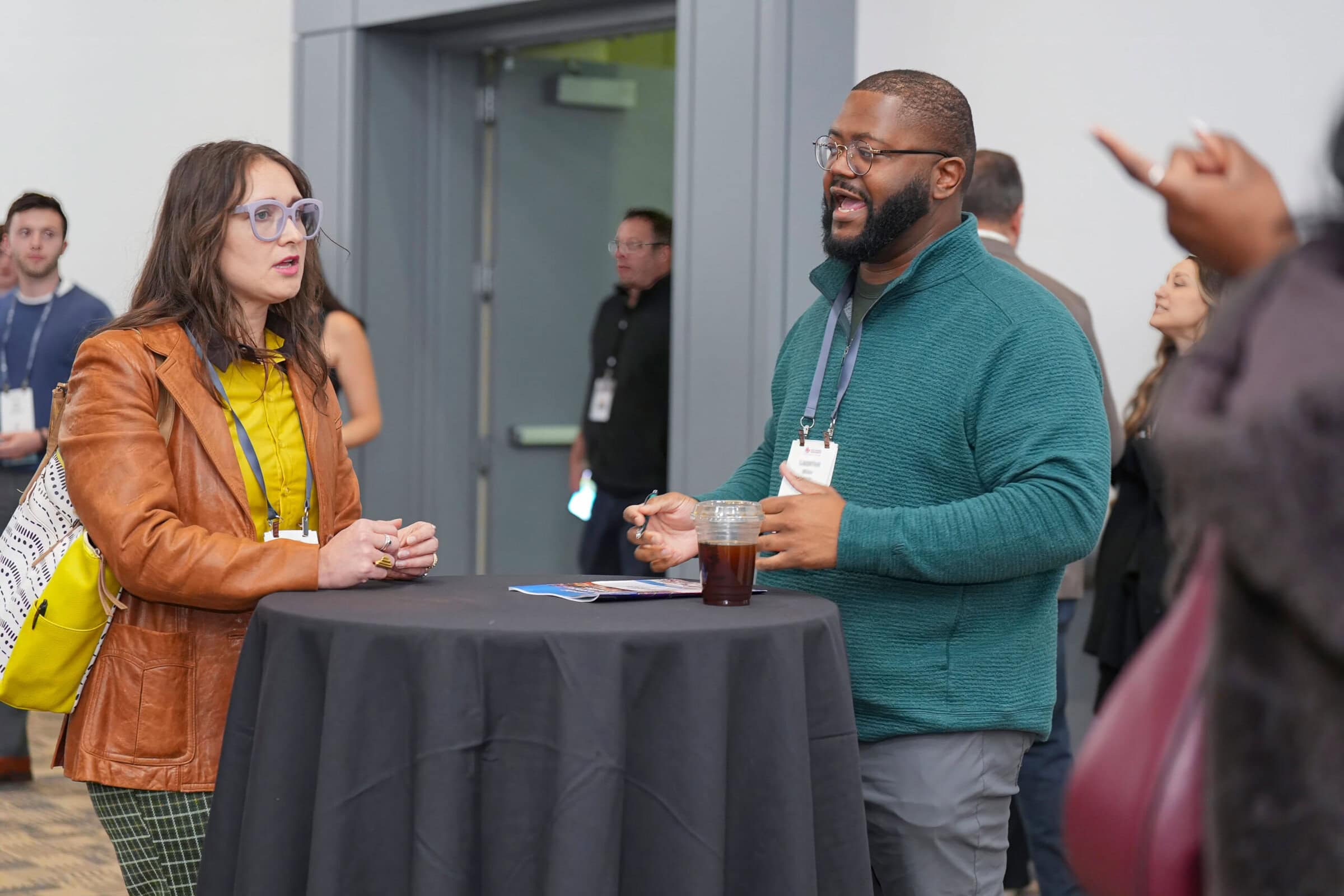 Two individuals discussing at a table in an event setting, with other attendees visible in the background.