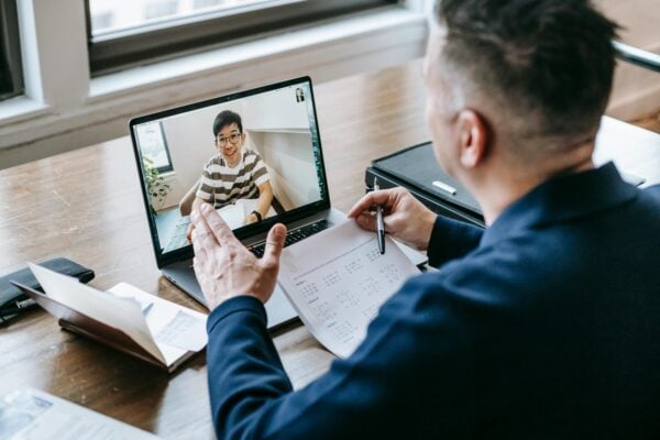 A man sits at a desk with a laptop, engaged in a video call displayed on the screen.