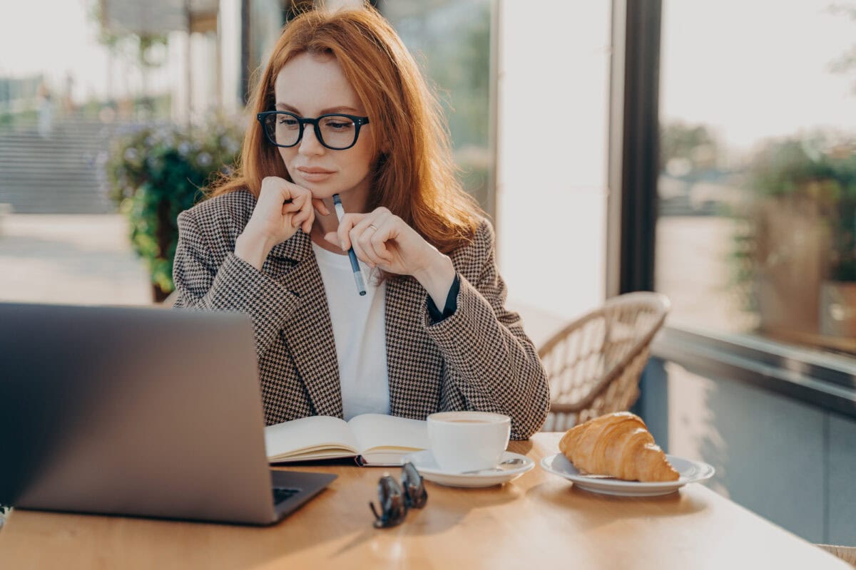 Serious businesswoman in formal wear works in cafeteria develops new startup project poses against coffee shop interior makes notes in notepad works remotely.