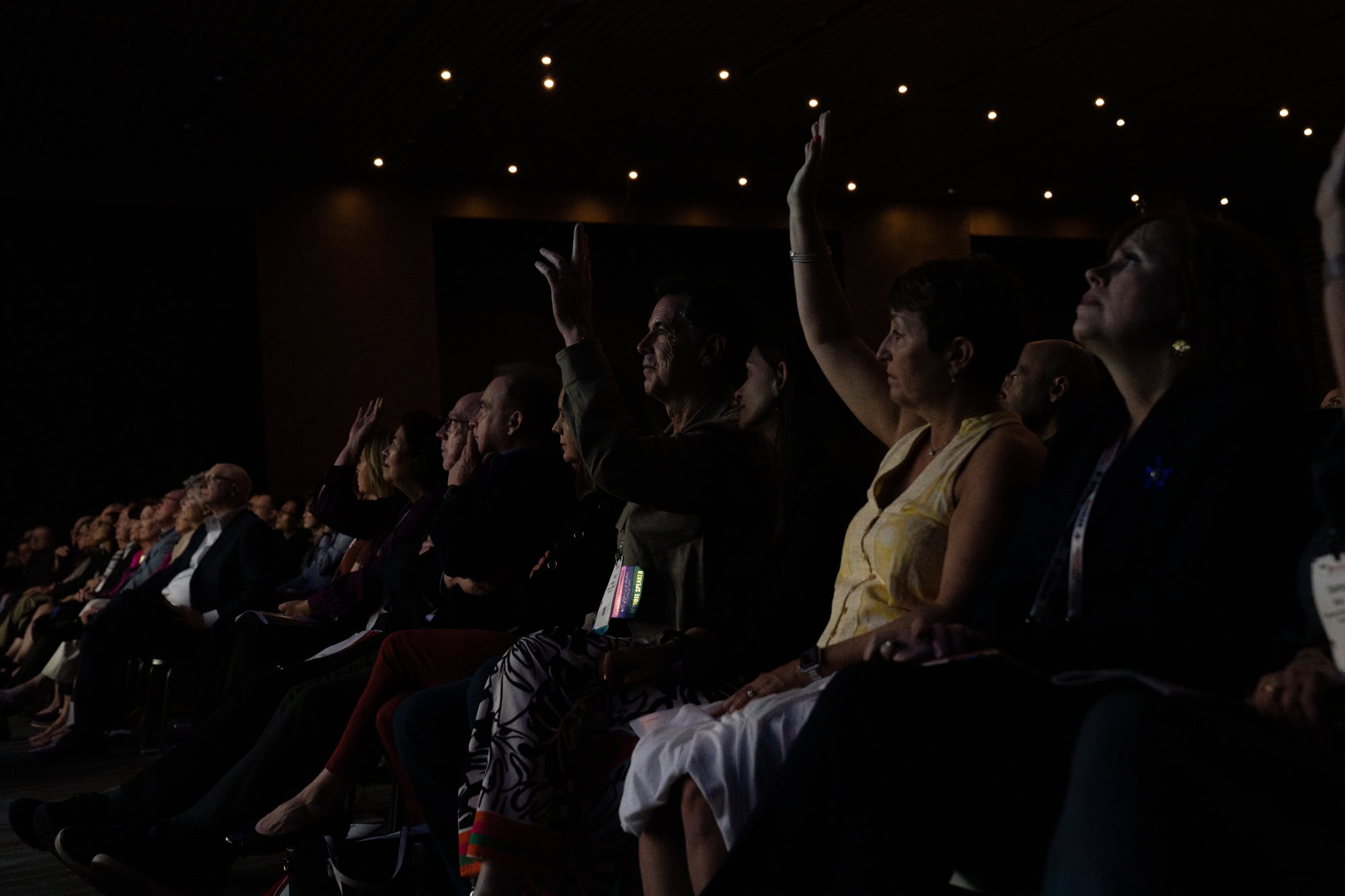 Audience members at a conference sit in a dimly lit auditorium, with several attendees raising their hands to participate during a presentation.