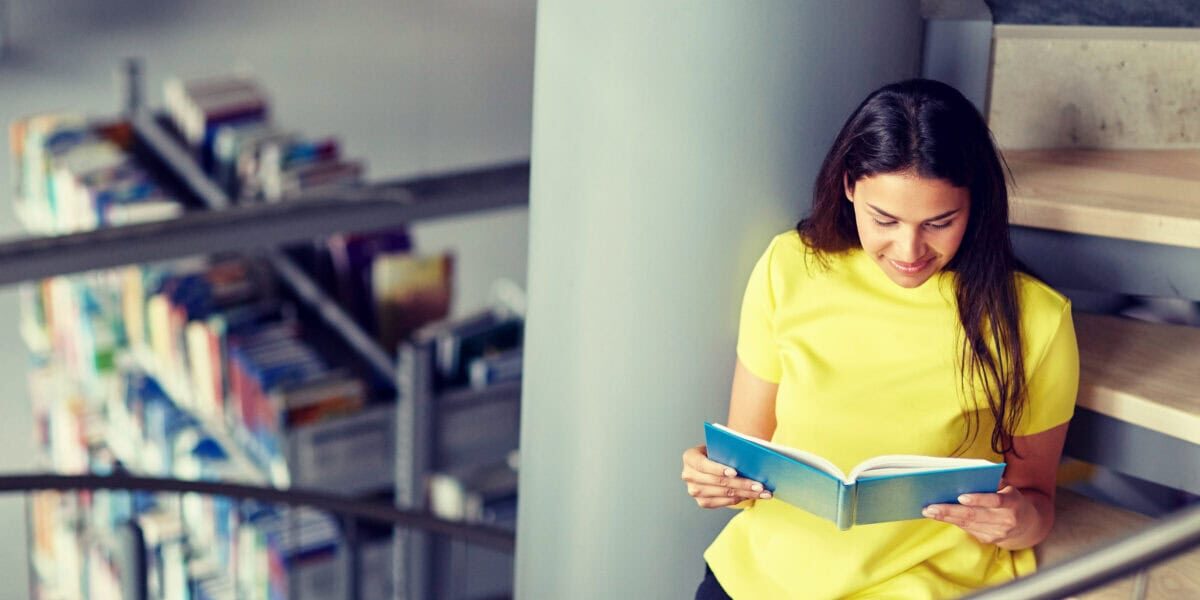 Smiling student girl, in a yellow blouse, reading book sitting on stairs at library