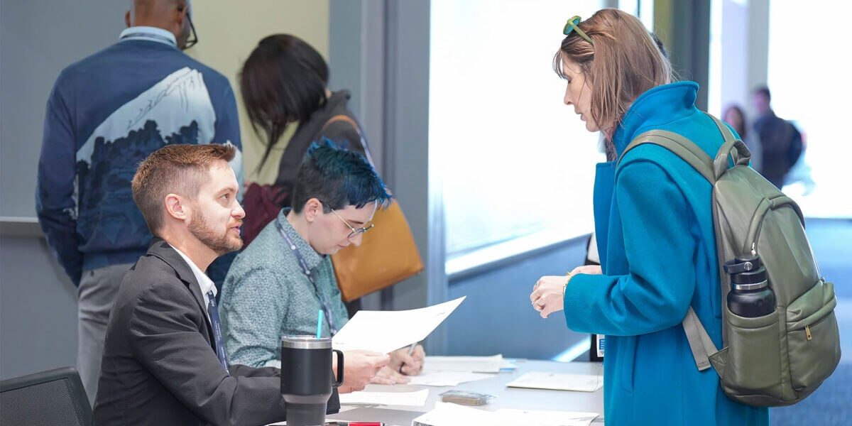 A man and woman engage in conversation at a check-in desk, surrounded by various company booths and attendees.
