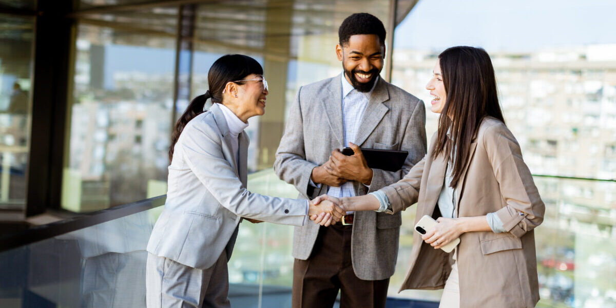 Two women and a man dressed in business attire engage in a warm handshake while smiling.