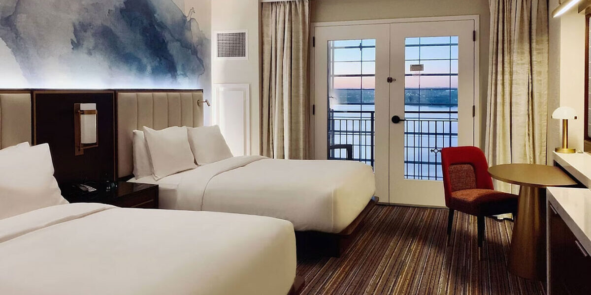 Modern hotel room with two beds, balcony, and scenic water view.