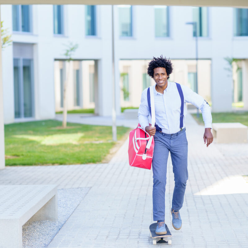 Smiling African American male entrepreneur with handbag riding skateboard along street in summer and looking at camera while commuting to work