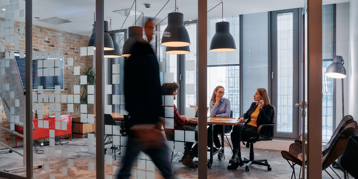 a group of young business professionals walk down the corridor next to their office, while their colleagues collaborate inside