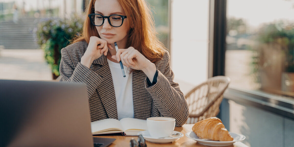 Serious businesswoman in formal wear works in cafeteria develops new startup project poses against coffee shop interior makes notes in notepad works remotely.