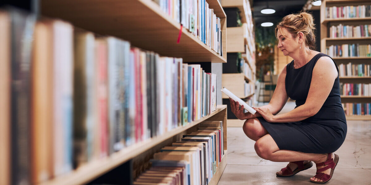 Woman in black dress, squatting down and choosing book in public library.