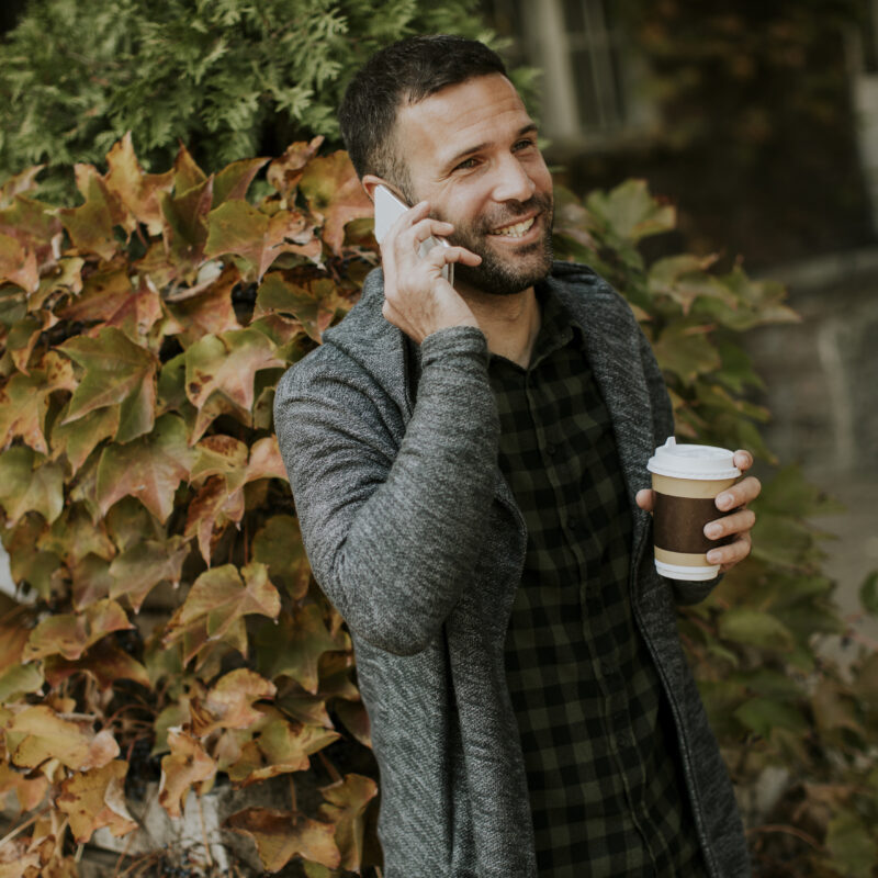 Handsome young man using mobile phone and holding takeaway coffee in the park