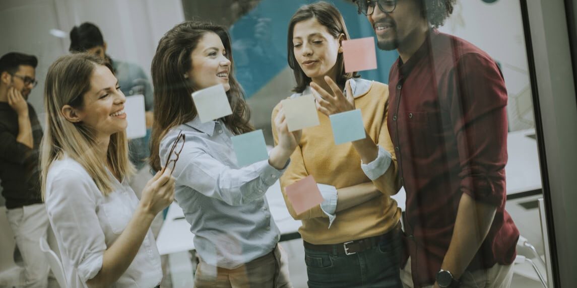 A group of people collaborates around a glass wall covered in colorful sticky notes, discussing ideas and strategies.
