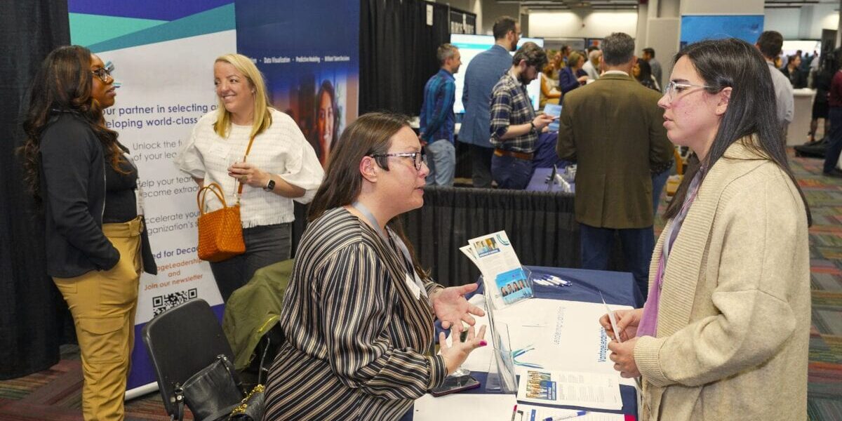 Two women engaged in conversation at a booth, surrounded by various company booths and attendees.