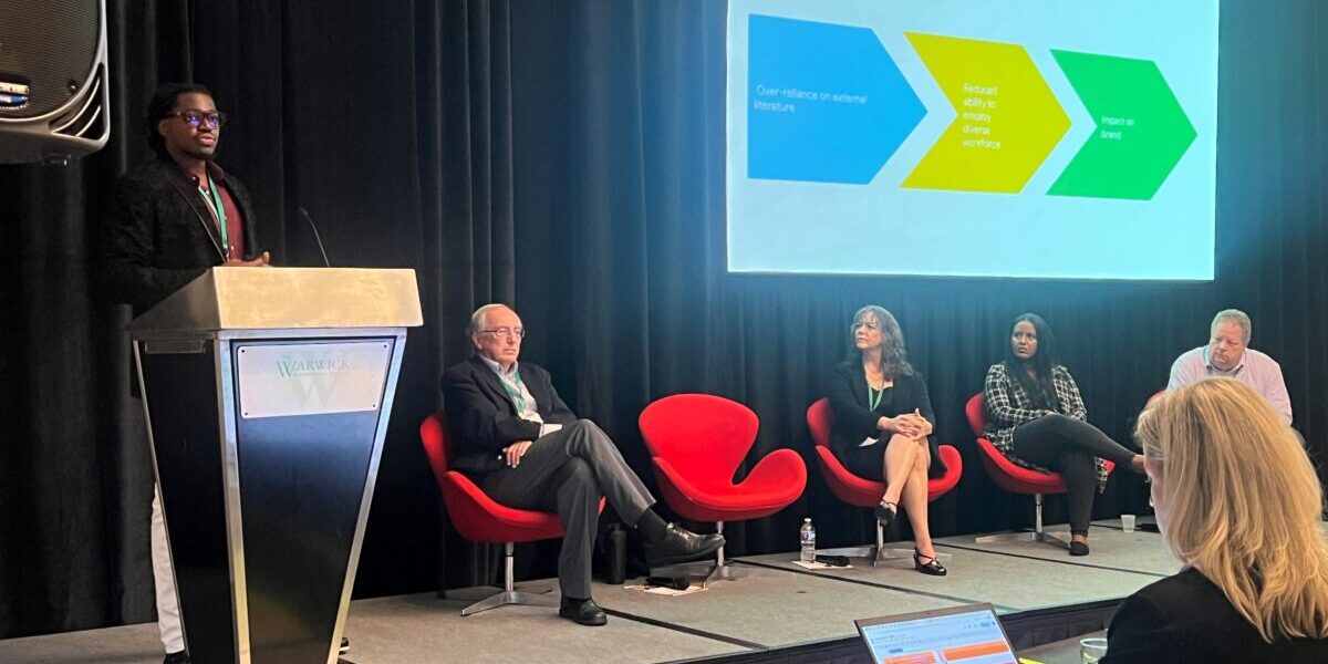 A group of people seated in chairs, on a stage, engaged in discussion. While a speaker is at a podium displaying information on a screen.