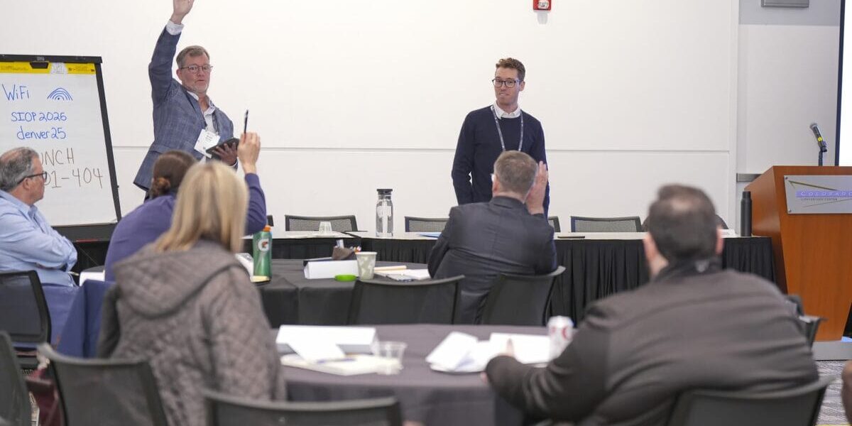 Attendees sitting at tables in a conference room, participating in a workshop. While speaker is talking in front of room with hand raised.