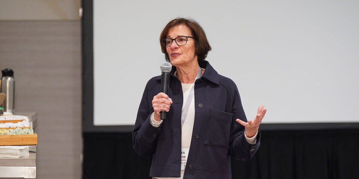 A woman with glasses addresses an audience, speaking into a microphone with an engaged expression.
