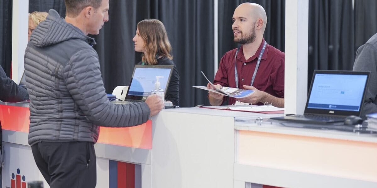 People standing at a booth with laptops and a man talking to someone.
