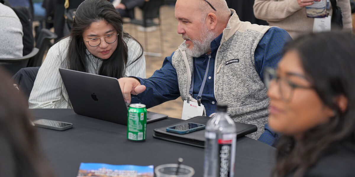 A man and woman sit at a table, focused on a laptop, engaged in discussion.