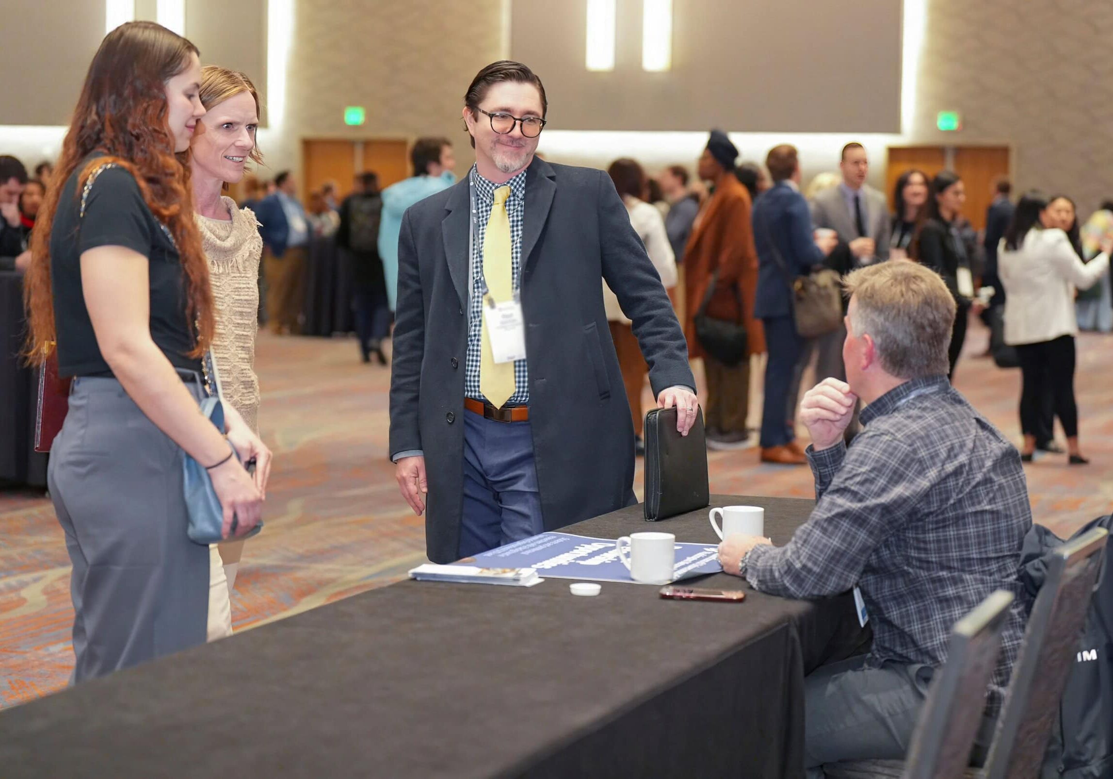 Four people conversing at a conference, a man sitting at a table while three are standing.