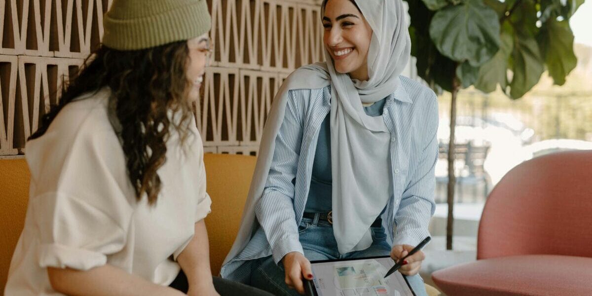 two women, one in a hijab and the other in a green beanie, sit on a couch next to each other talking