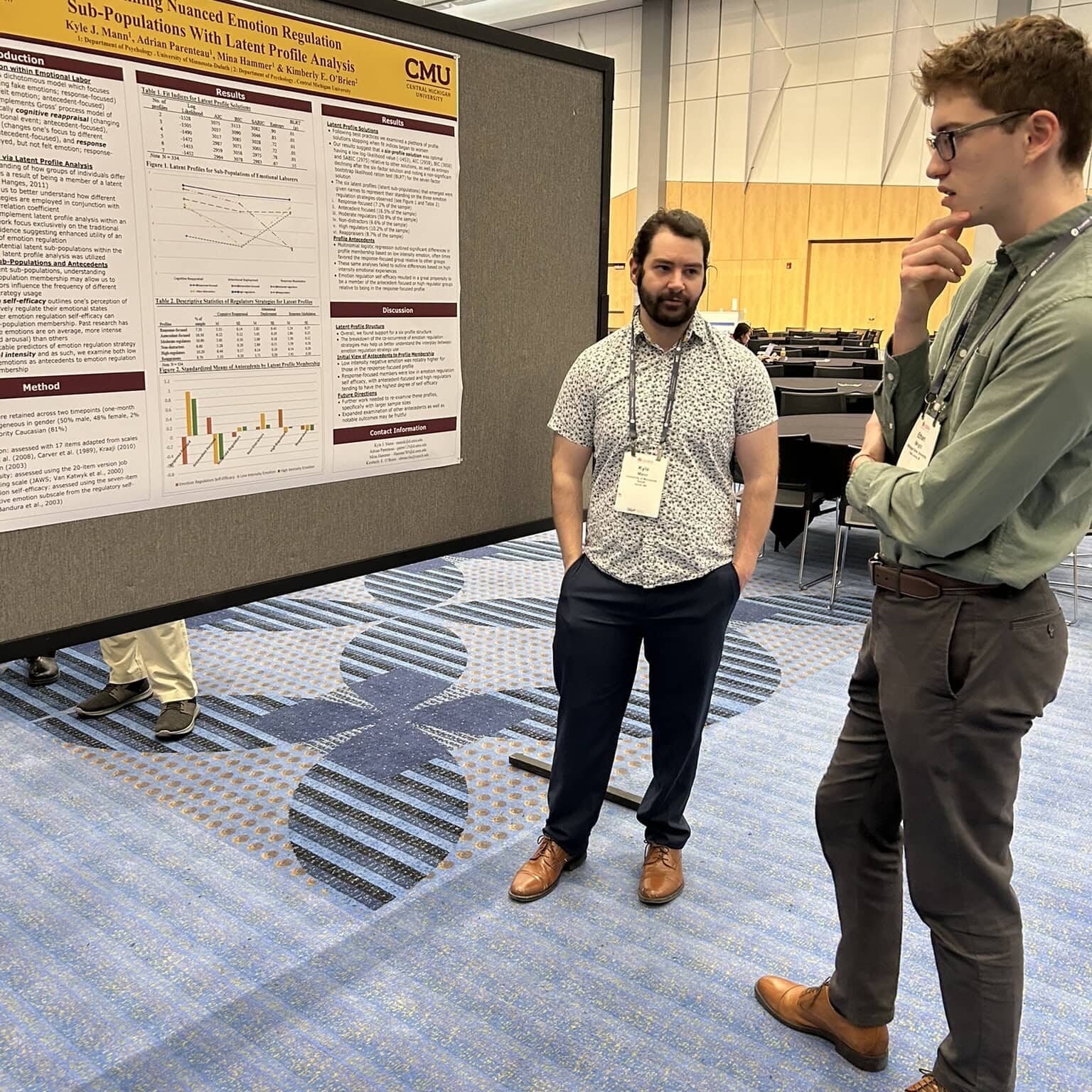 Two men stand in front of a colorful poster, engaged in conversation and looking at the displayed information.
