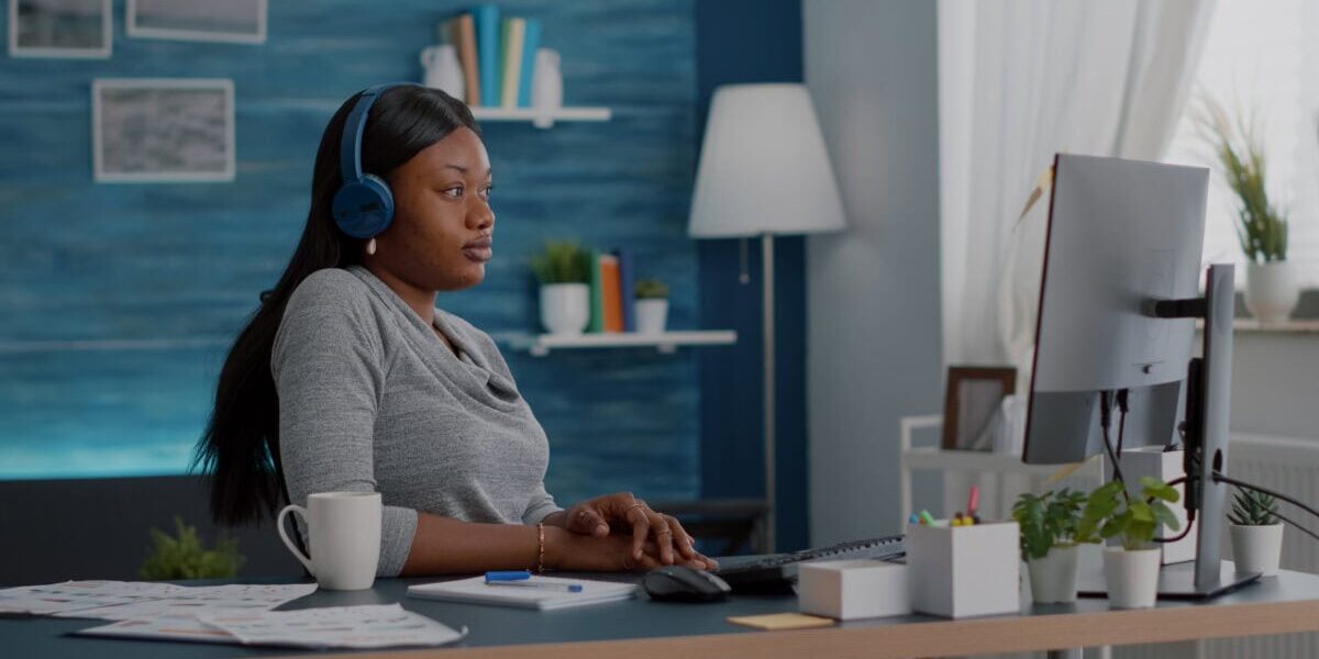Student with black skin having headphone puts listening online univeristy course using elearning platform sitting at desk in living room. African american woman working remote from home